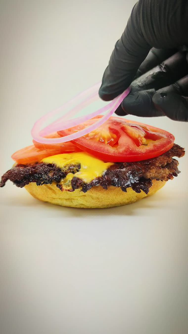 Hamburguesa de Only Burger armándose: carne, queso y tomate sobre el pan, mientras una mano agrega el aro de cebolla