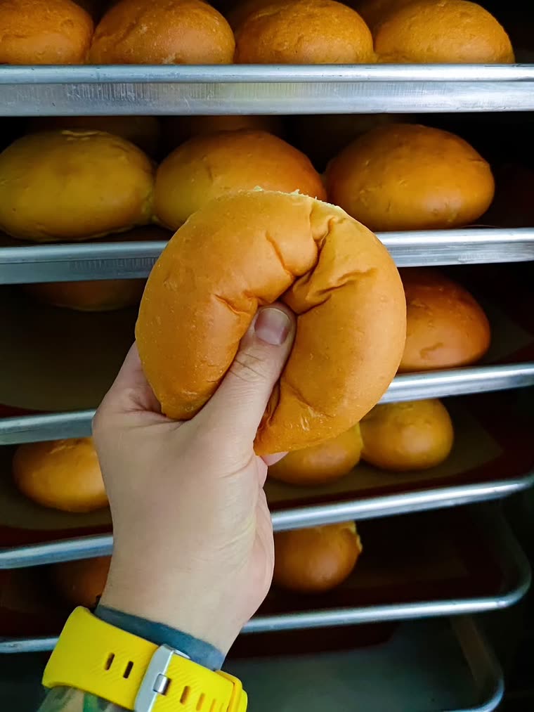 Pan de papa recién horneado, dorado y liso, sostenido frente a las bandejas del carro
